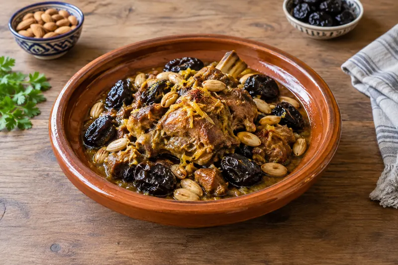 A shallow earthenware dish of dark lamb stew topped with glossy prunes, whole golden almonds and a scattering of sesame seeds