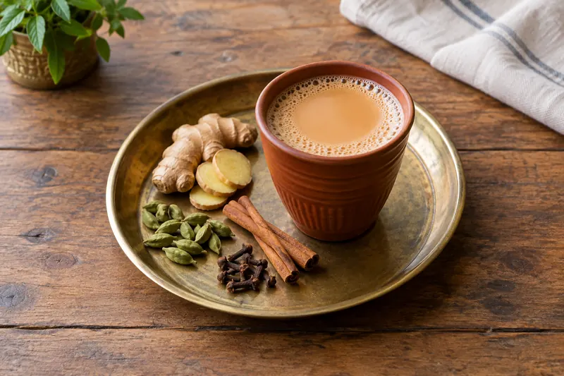 Two small glasses of milky spiced tea beside a pan, ginger, cardamom and cinnamon